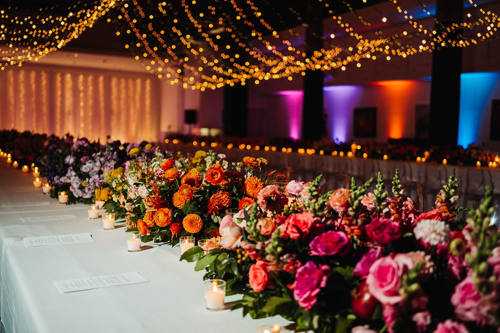 Decorative floral arrangements on a long table with colorful lighting in the background